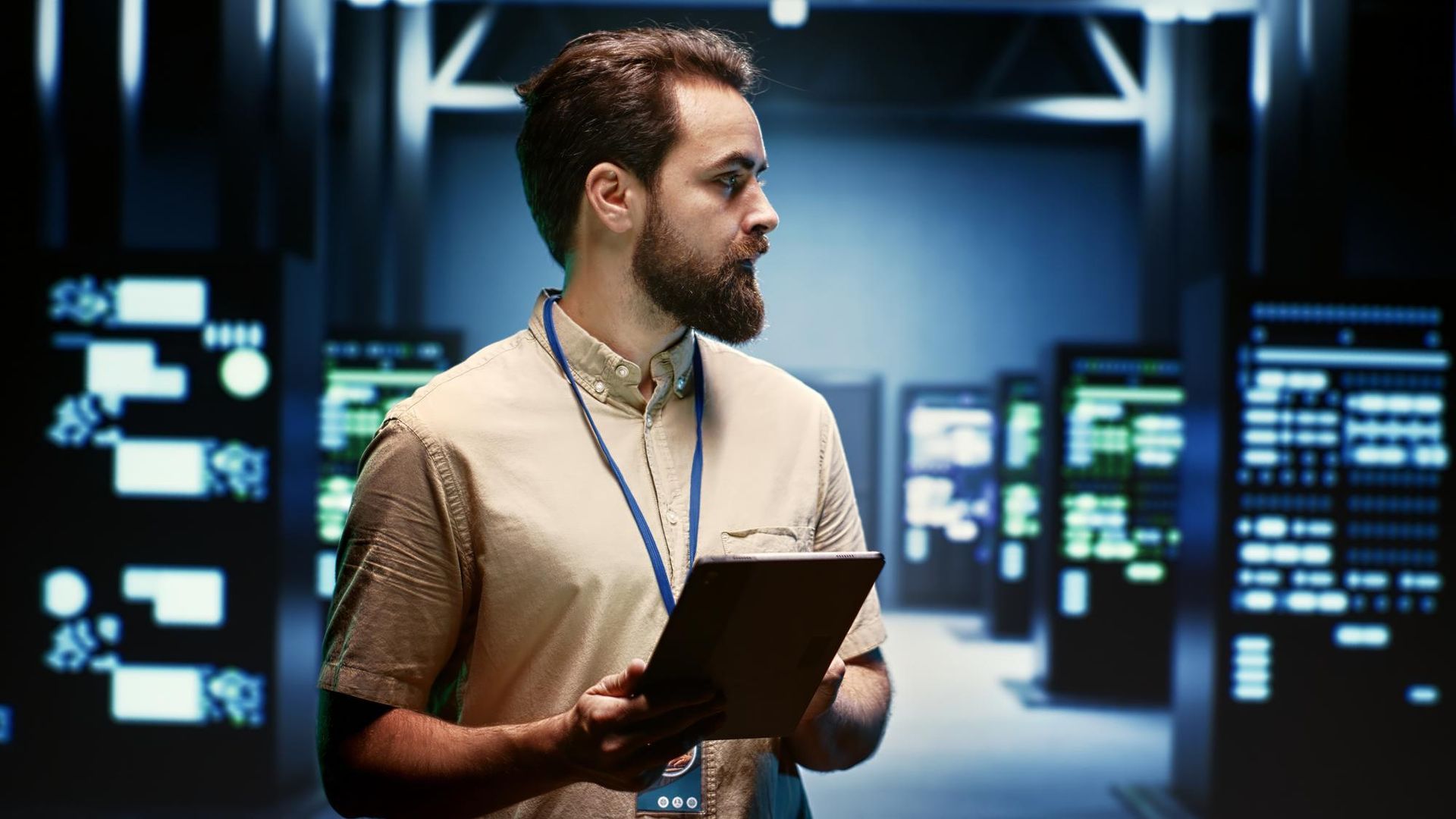 A man with a beard is holding a tablet in a server room.