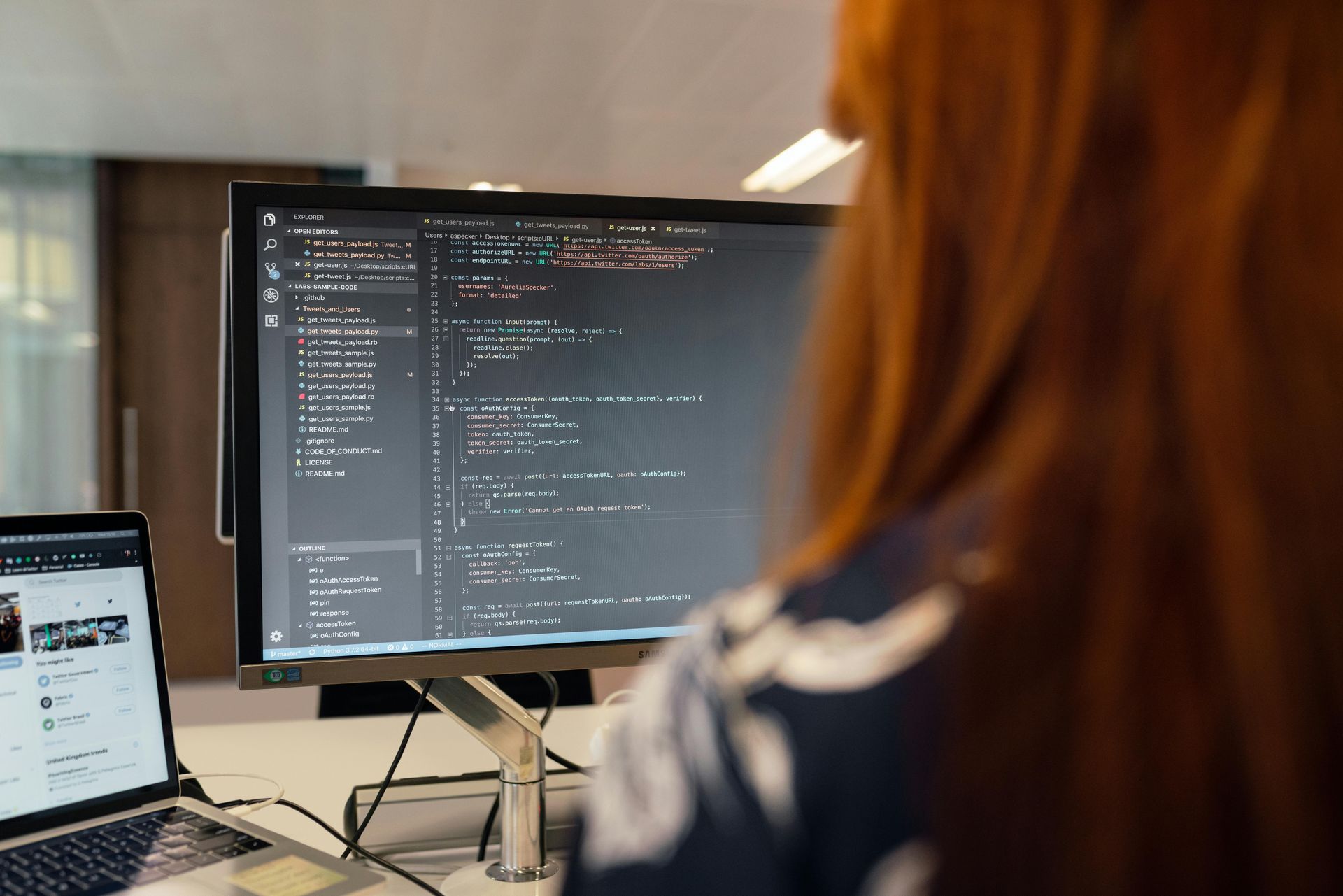 Woman with red hair, coding at a computer. A laptop is visible on the desk. The setting is an office.