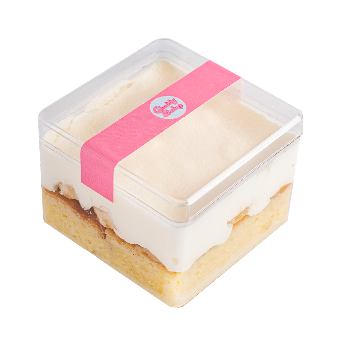 A square dessert in a clear plastic container, layered with cake, cream, and sauce, sealed with a pink label.
