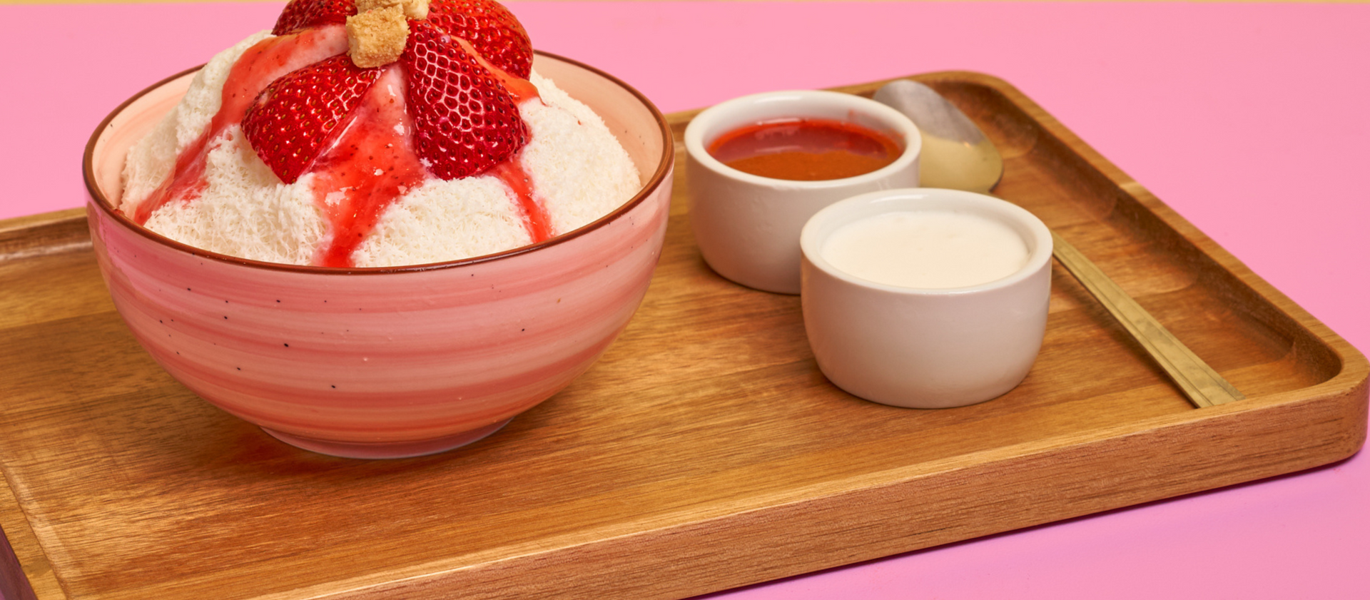 Strawberry shaved ice dessert in a pink bowl on a wooden tray with sauces on the side.