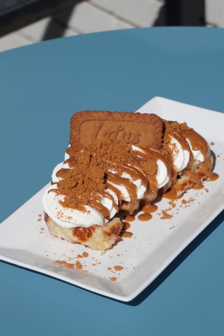 A dessert on a white rectangular plate: cinnamon roll slices topped with whipped cream, Biscoff cookie crumbles, and drizzled sauce, with a Biscoff cookie on top; blue table background.