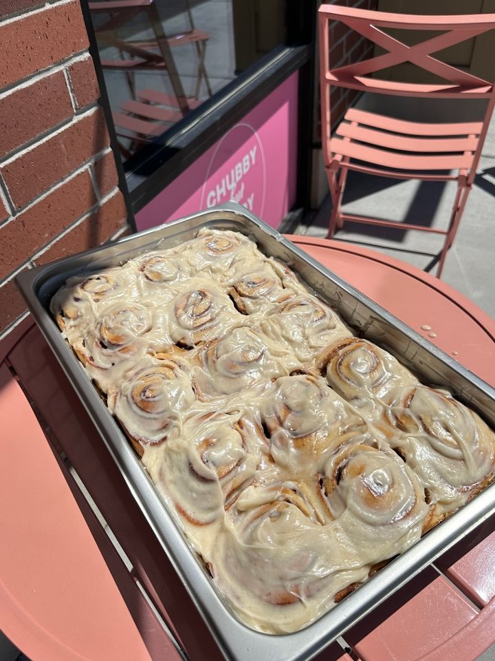 A tray of frosted cinnamon rolls sits on a pink table outside a bakery.