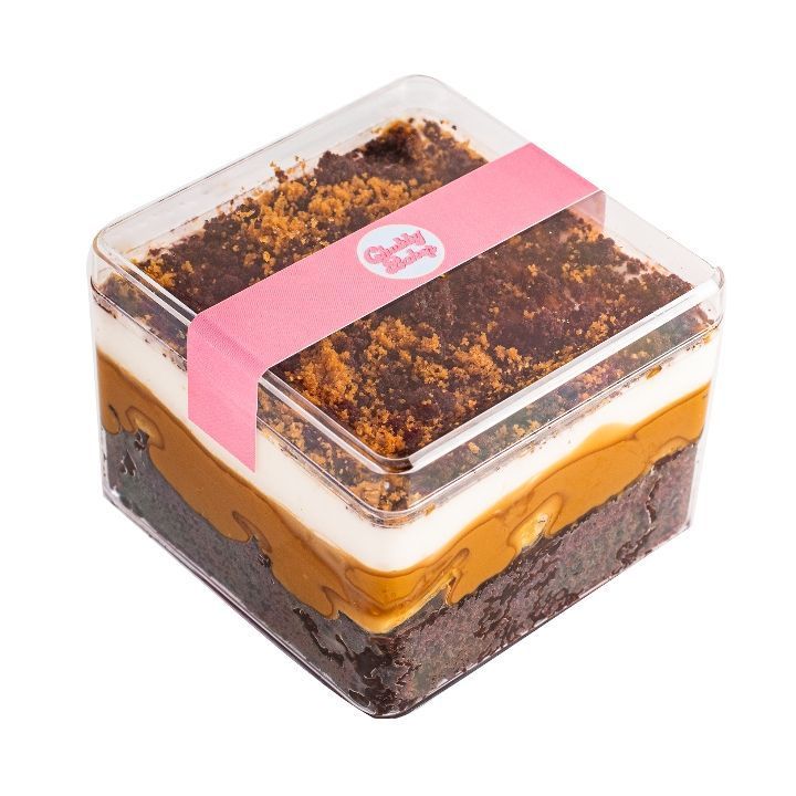Square dessert container with layers: brownie, caramel, white cream, and crumbled topping. Pink label reads 