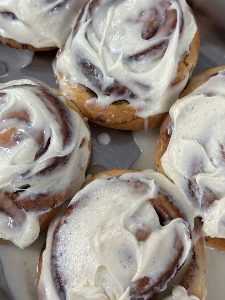 Close-up of frosted cinnamon rolls in a metal baking tray. Creamy white frosting generously covers each roll.