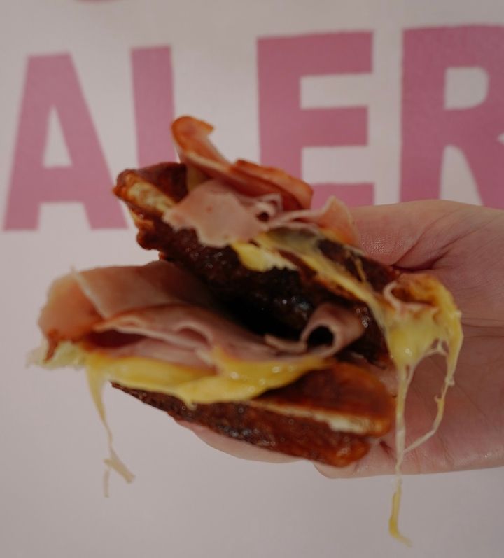 Hand holding a grilled sandwich overflowing with melted cheese and ham; a pink background is visible.