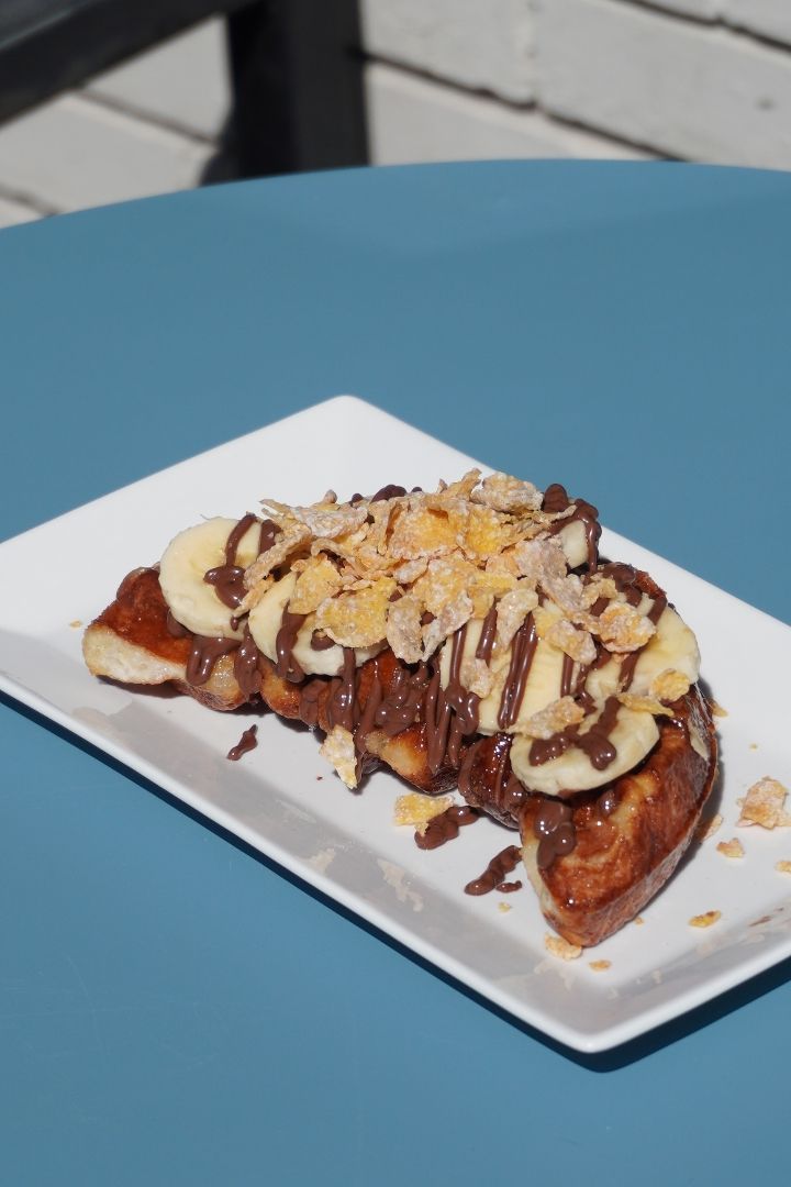 Croissant waffle topped with chocolate sauce, banana slices, and almond flakes on a white rectangular plate on a blue table.