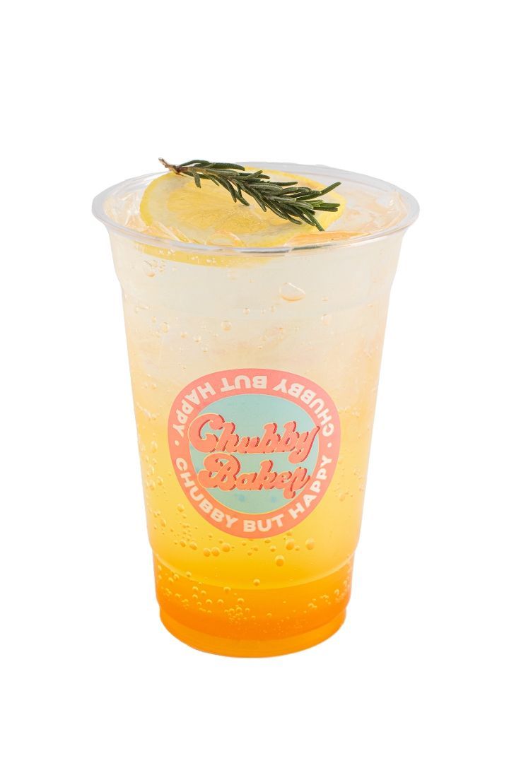 A refreshing yellow and white drink in a clear plastic cup, garnished with a lemon slice and rosemary. The cup has a 