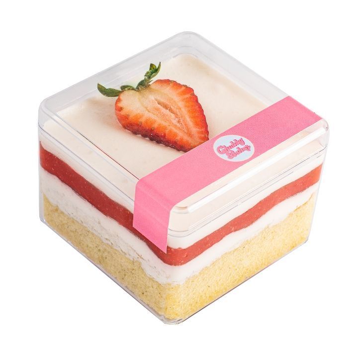 A layered strawberry shortcake in a clear square container, topped with a halved strawberry. Pink label wraps the side.