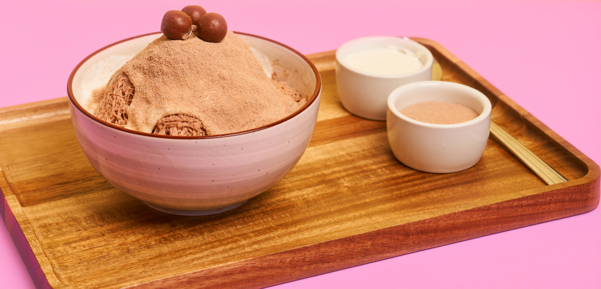 Chocolate shaved ice dessert in a bowl, served on a wooden tray with two small bowls of toppings. Pink background.