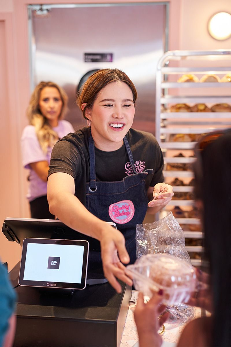 A smiling bakery worker in a black shirt and apron hands a bag of treats to a customer near a cash register; a woman waits in the background.