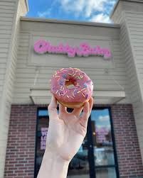 A hand holding a pink donut with sprinkles in front of a bakery called 
