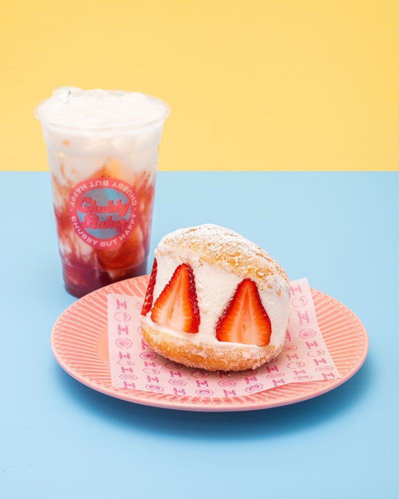 Strawberry donut with cream and fresh strawberries, plus a strawberry drink, on pink plate against blue and yellow backdrop.