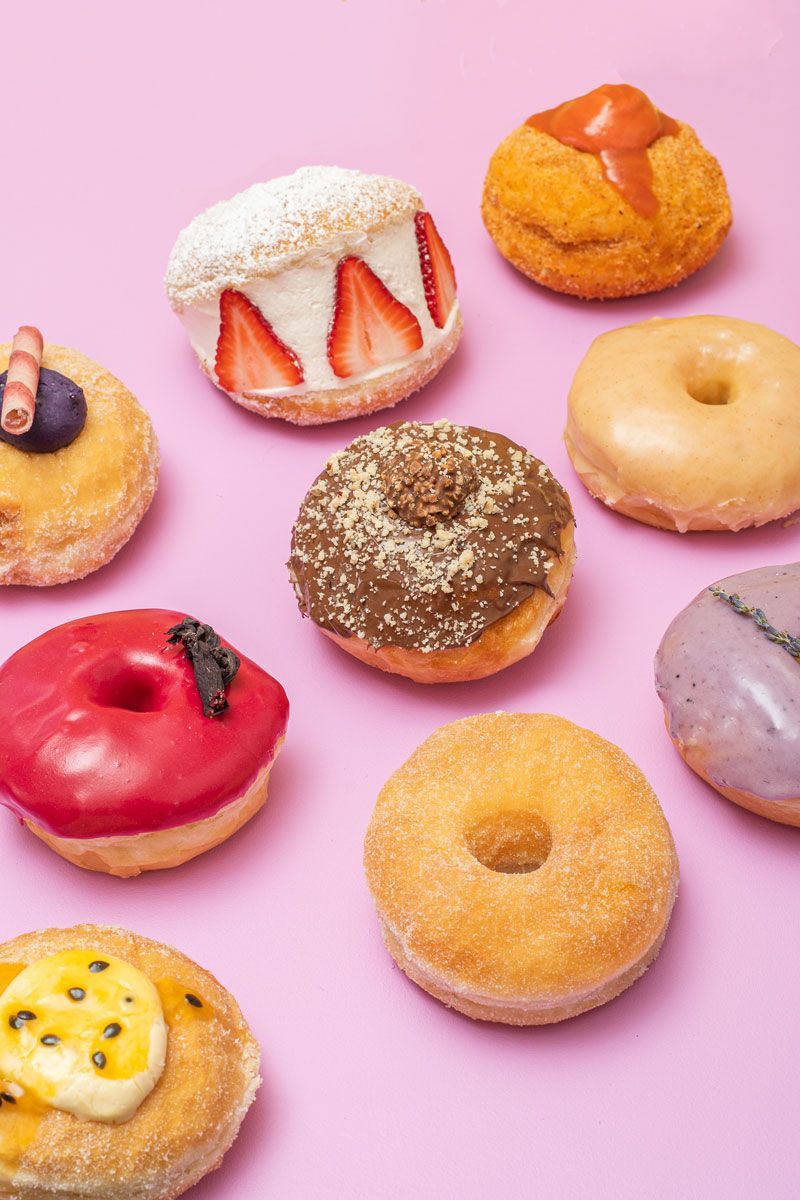 Assortment of colorful, glazed donuts on a pink background, including flavors like strawberry, chocolate, and passionfruit.
