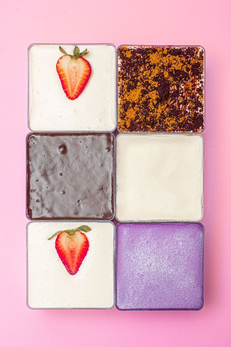 Six square desserts in clear containers, arranged on a pink background. The desserts have different toppings and colors, including strawberries, brown powder, and purple glitter.