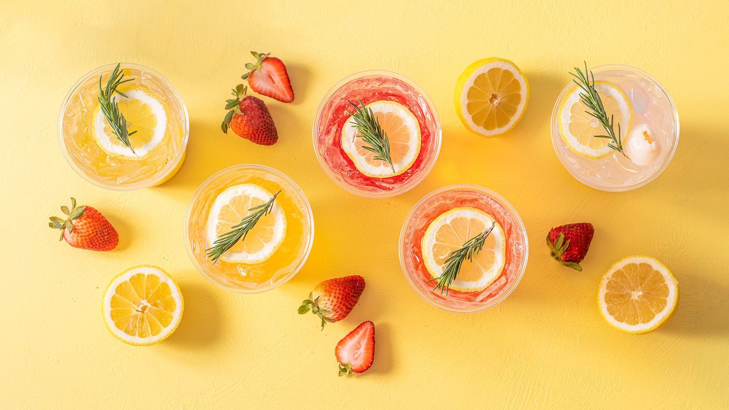 Cocktails with lemon slices, rosemary, and strawberries on a yellow surface.