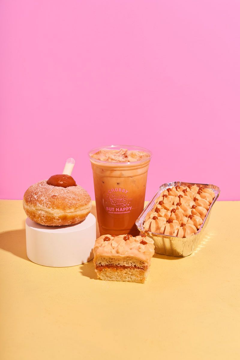 Donut with caramel topping, iced drink, cake slice, and baked dessert on a yellow surface against a pink backdrop.