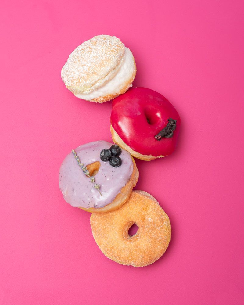 Four colorful donuts on a pink background. Frostings include white, red, lavender, and sugar.