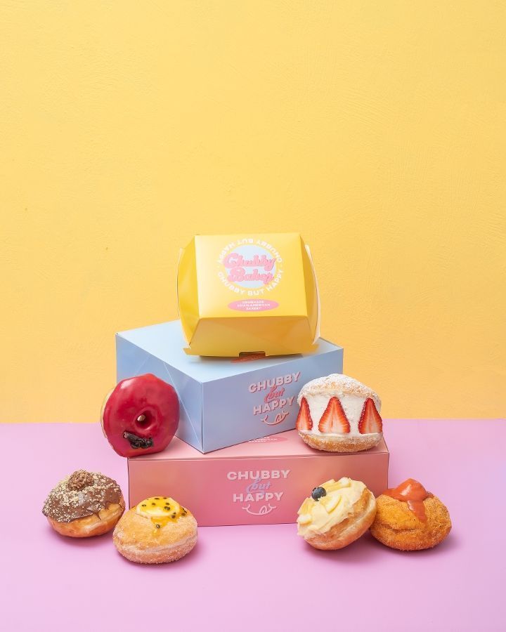 Chubby Happy pastries arranged on pink and blue boxes against a yellow backdrop. Includes a box, doughnuts, and cream-filled treats.