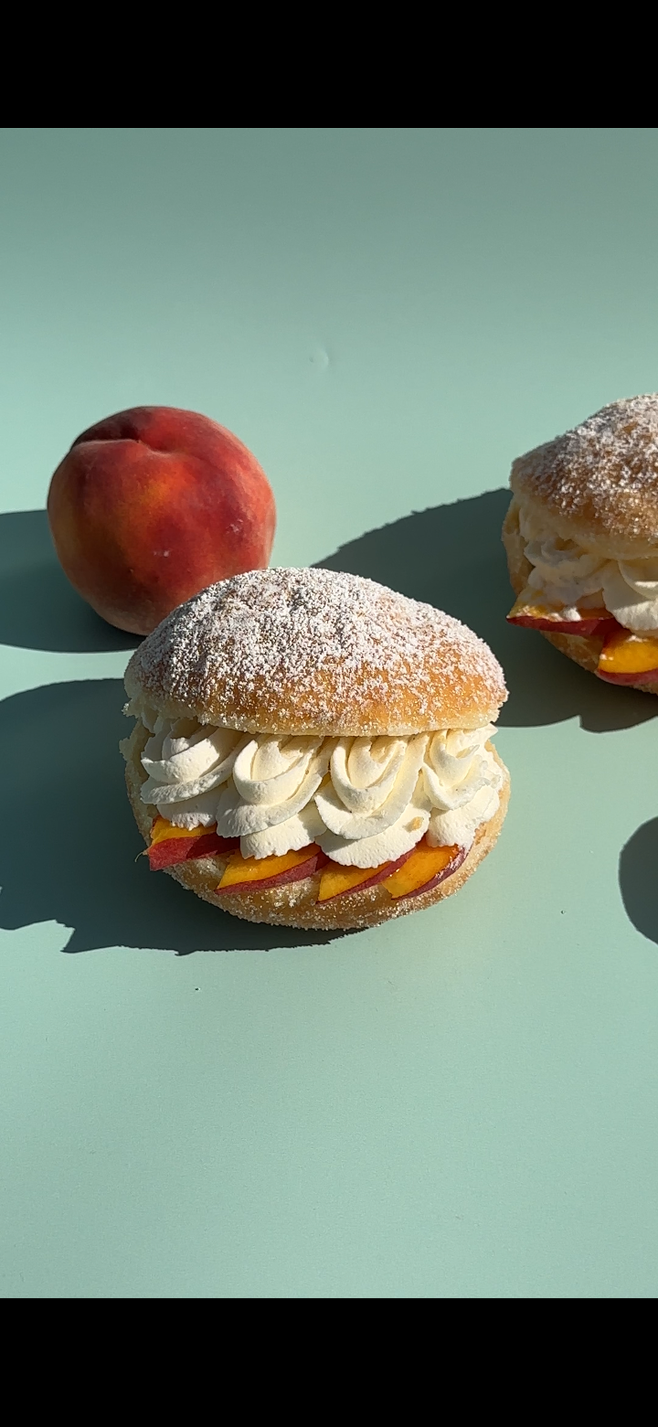 A peach biscuit sandwich filled with whipped cream and peach slices, dusted with powdered sugar, next to a whole peach.