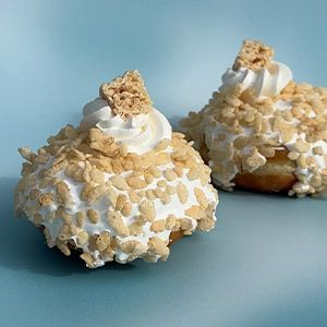 Two glazed pastries with nuts on top, sit on a blue-green surface with spilled glaze and scattered nuts, side-lit by sunlight.