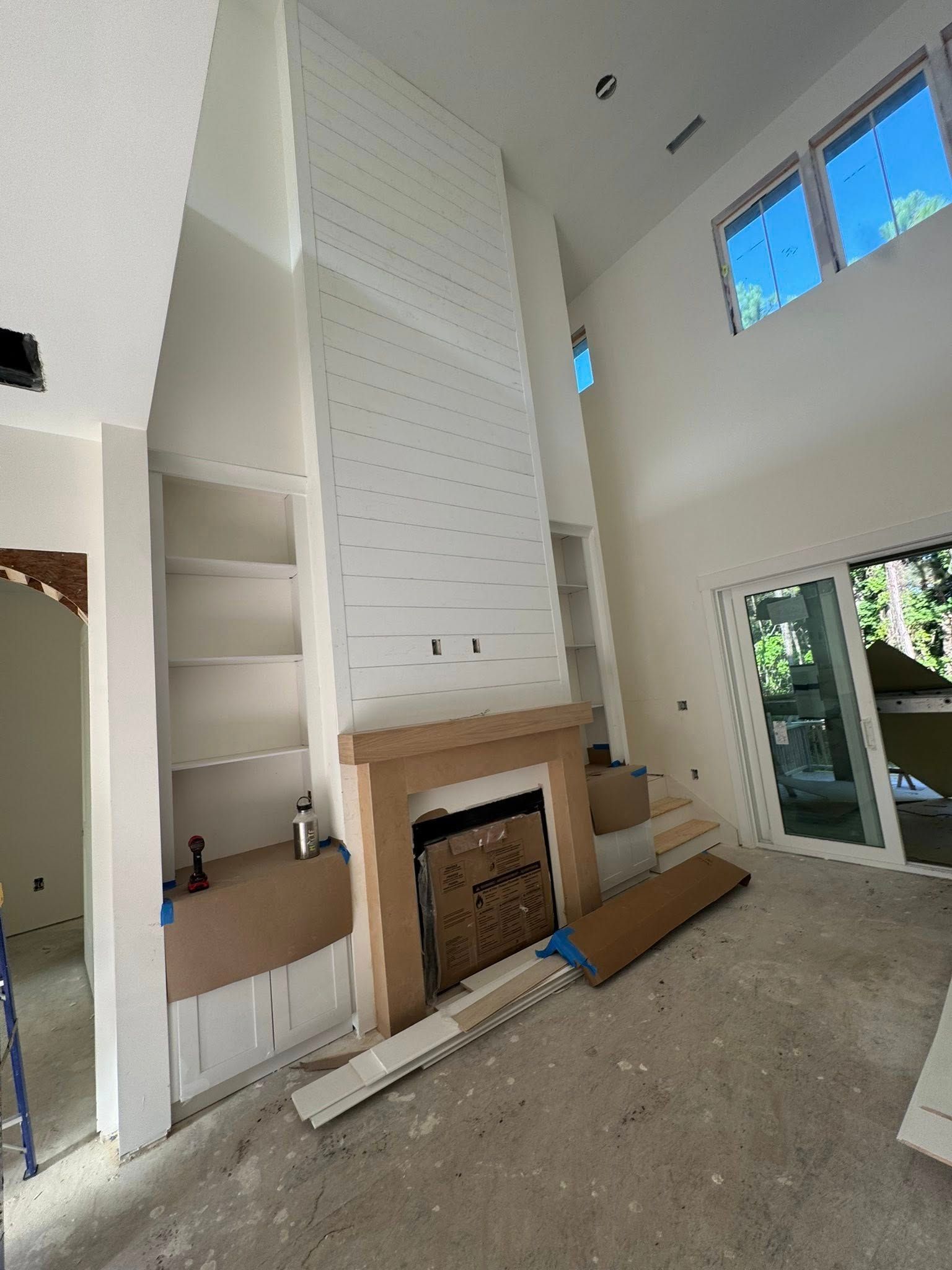 Interior of a room with a tall, white fireplace, built-in shelving, and large windows. Construction in progress.