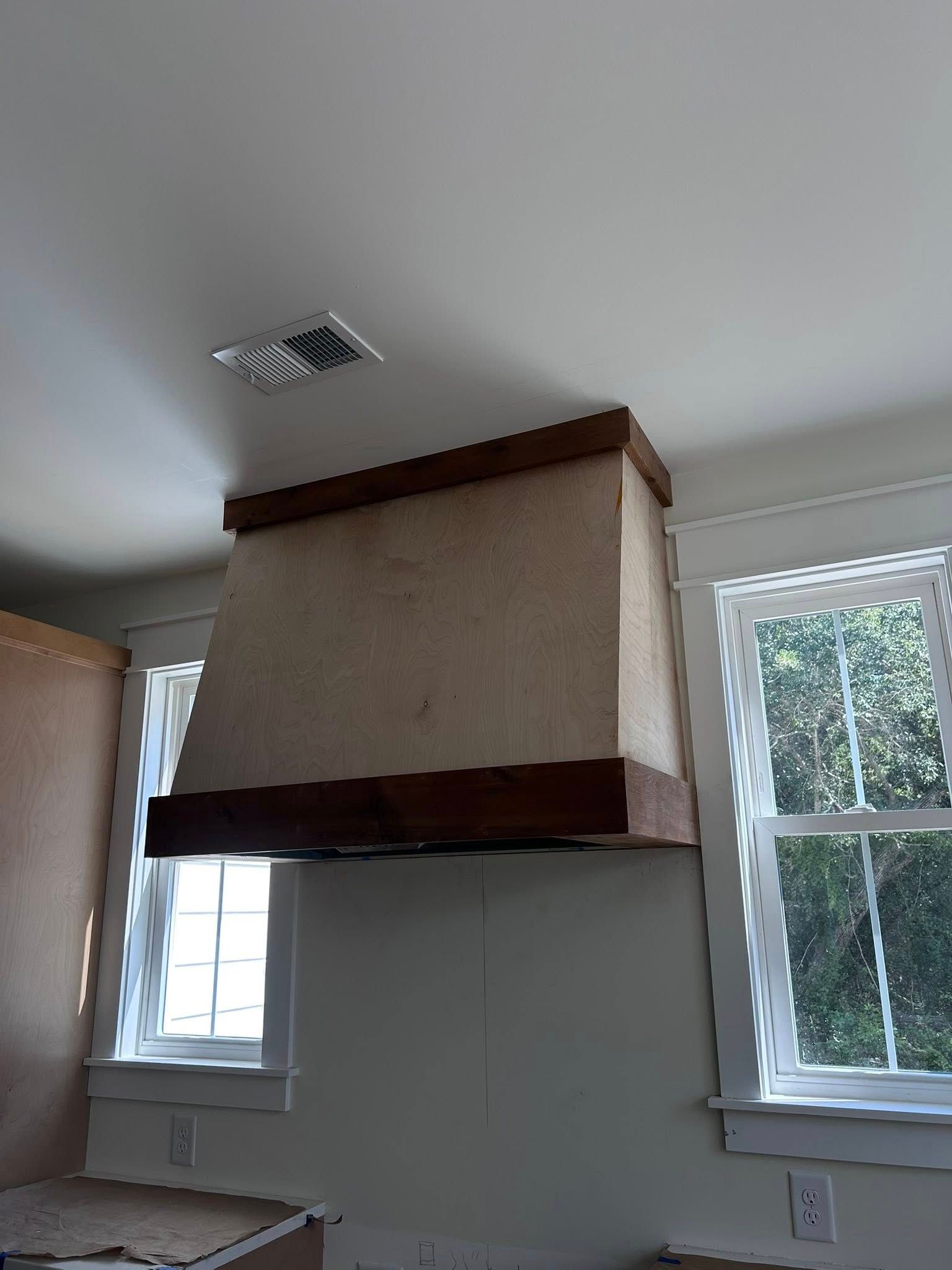 A kitchen range hood, tan fabric with wood trim, centered between two windows.