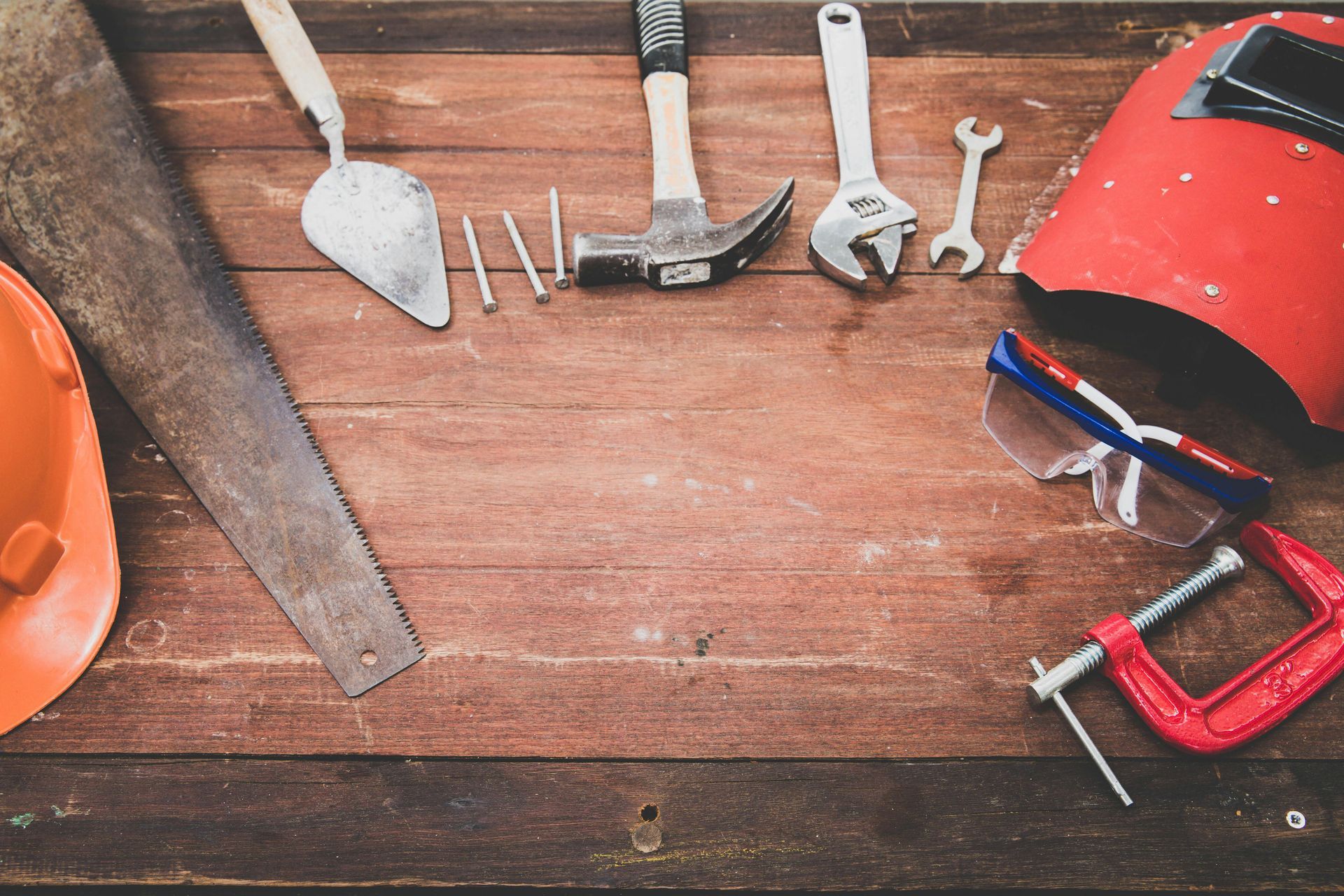 Tools laid out on a wooden surface: saw, trowel, nails, hammer, wrench, safety glasses, welding mask, clamp, and hard hat.