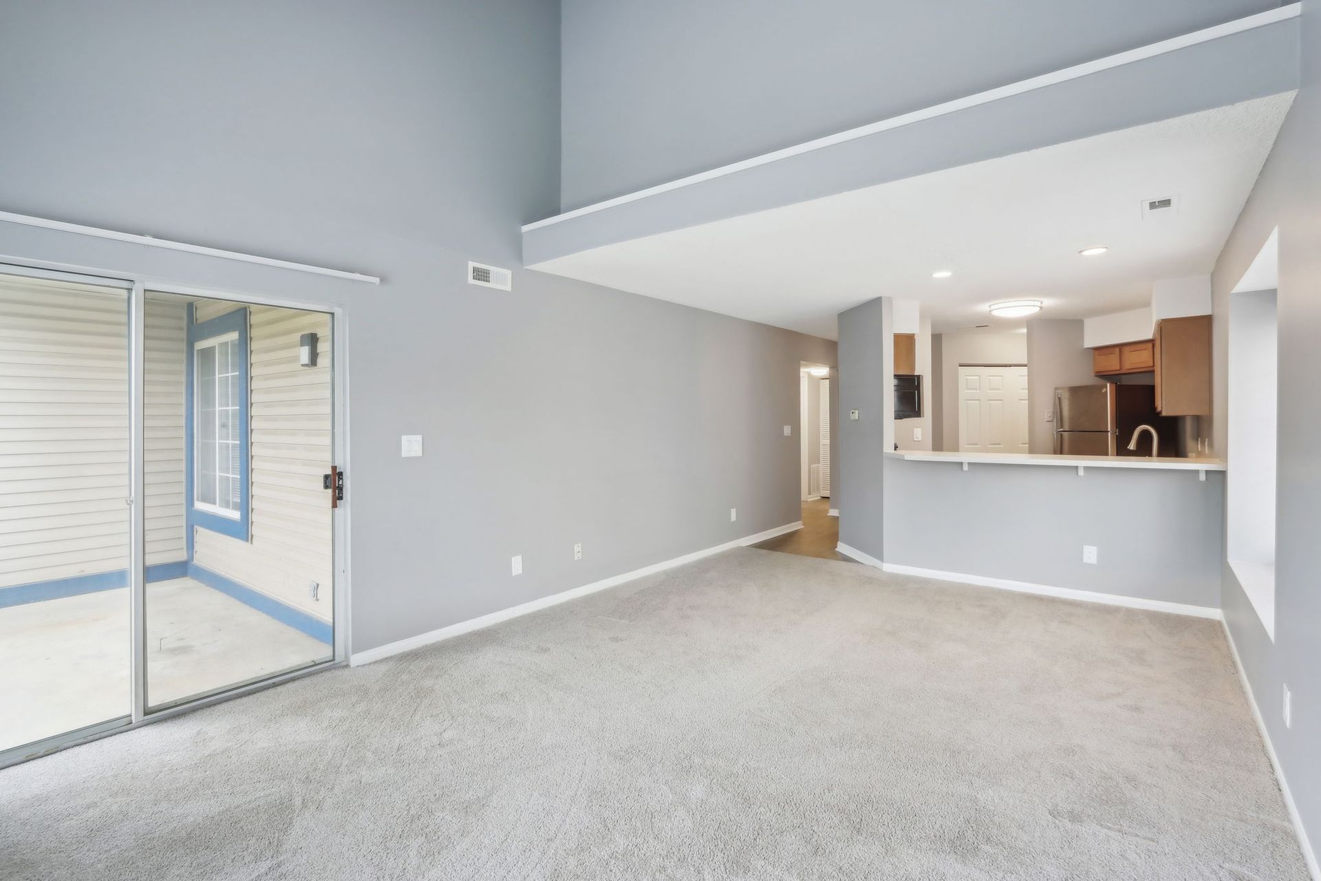 Empty living room with grey walls, carpet, and a partial view of a kitchen. Sliding glass door to a patio.