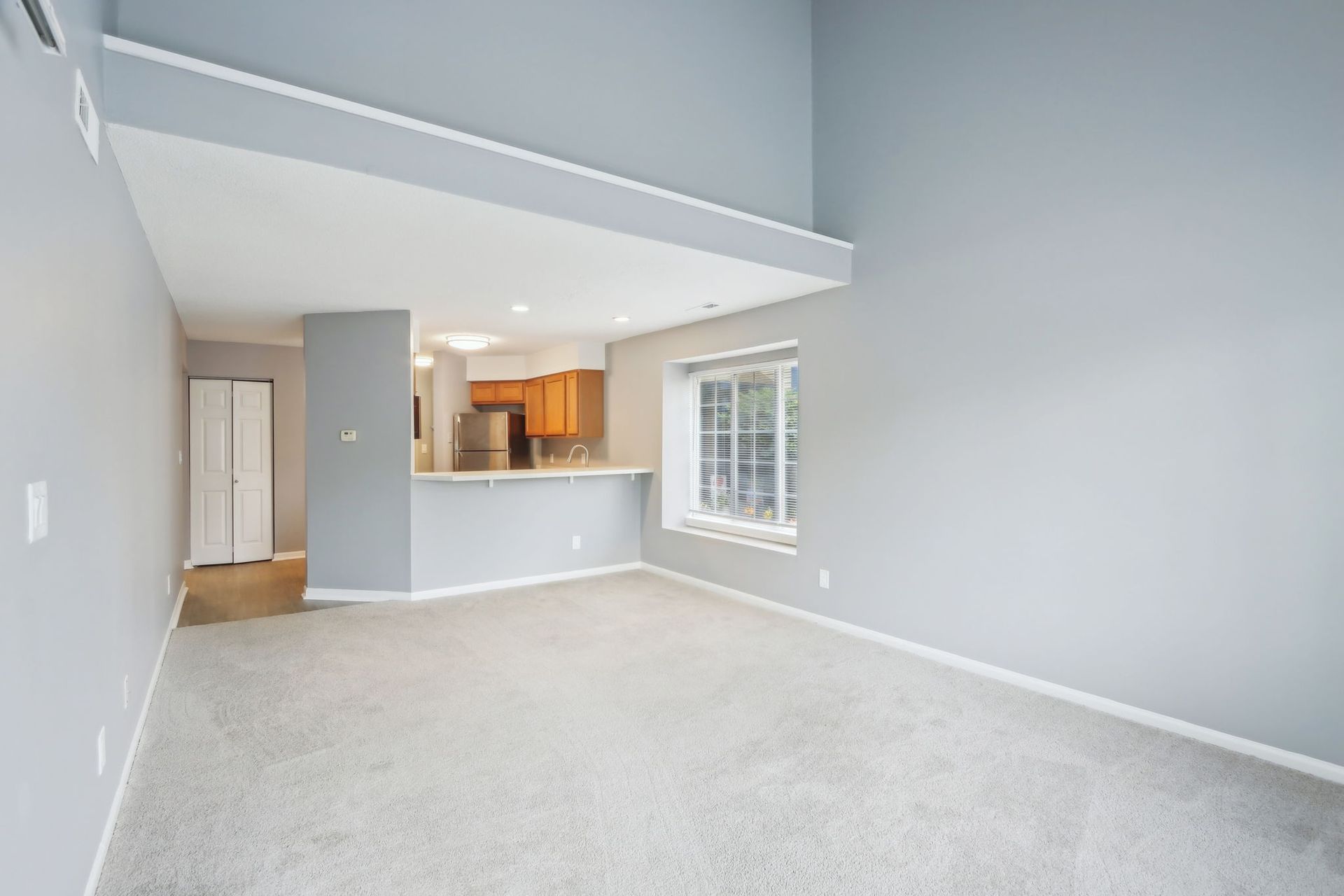 Empty living room with gray walls, beige carpet, and a view into the kitchen.