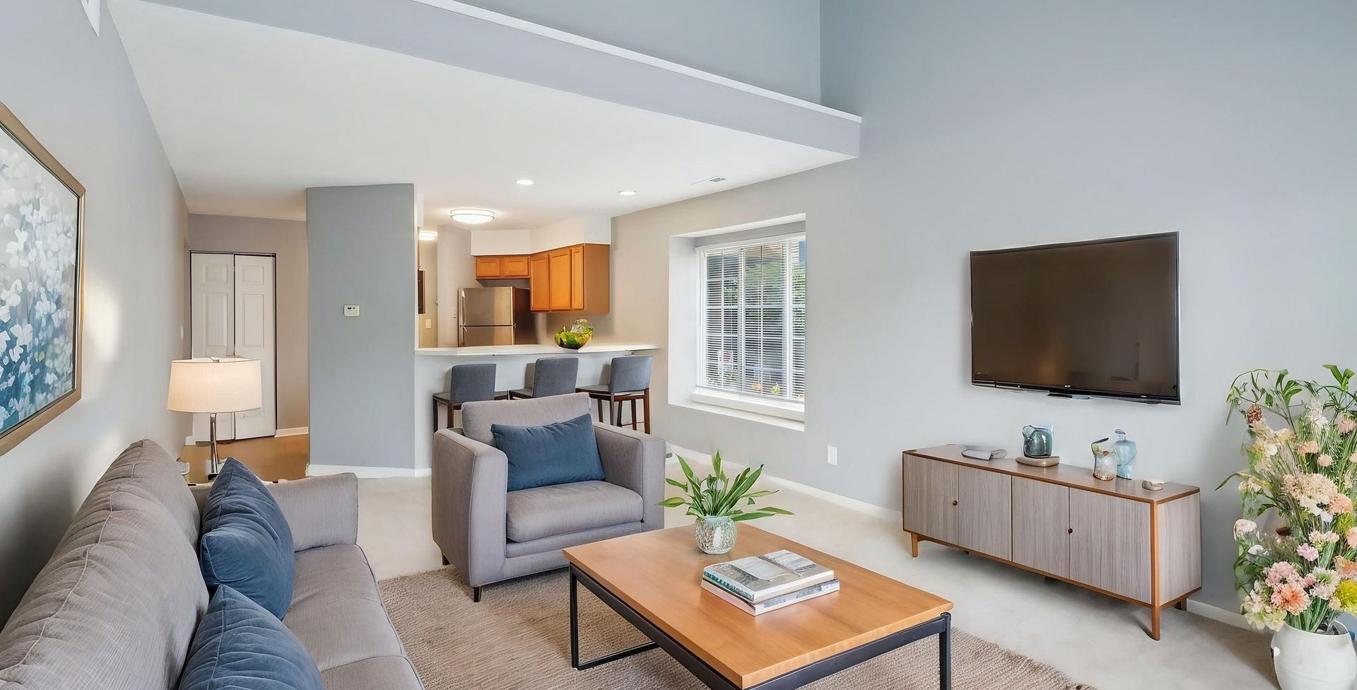 Cozy living room with a sofa, armchair, and TV. Light blue walls and a view into the kitchen.