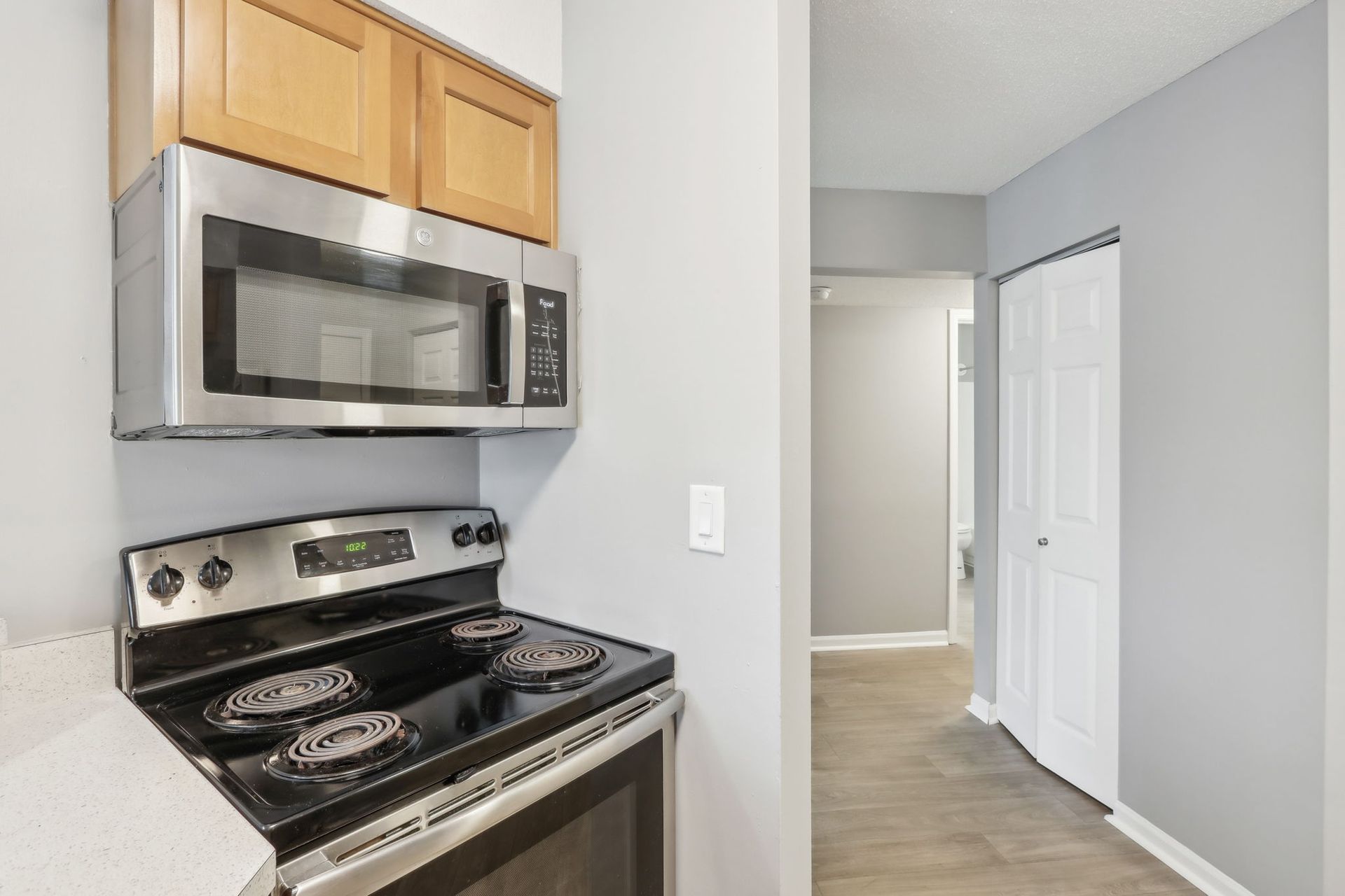 Kitchen with stainless steel appliances: microwave, stove, and light wood cabinets. Gray walls and hallway with white door.
