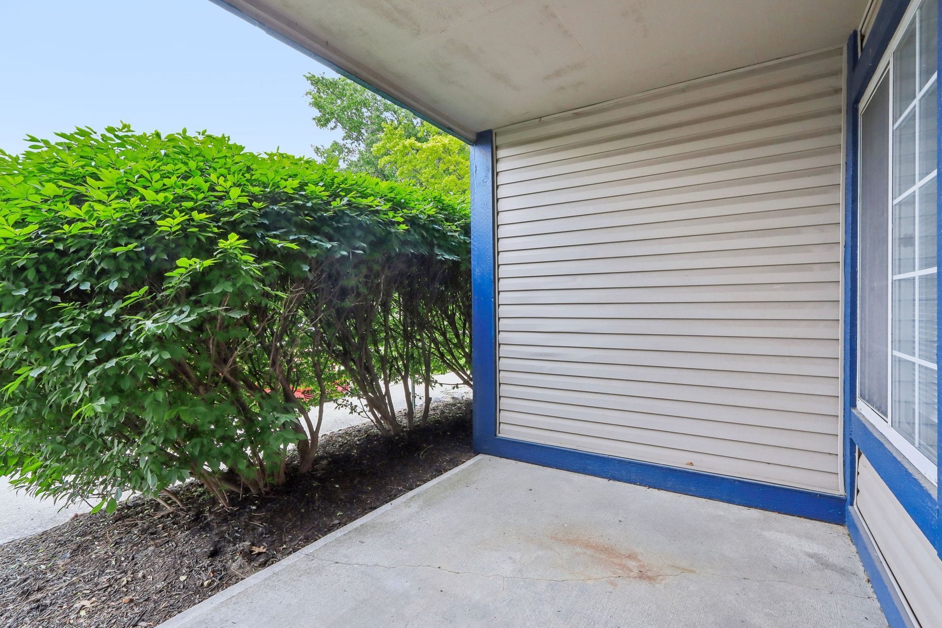 Covered outdoor porch with a large green bush, white siding, and blue trim.