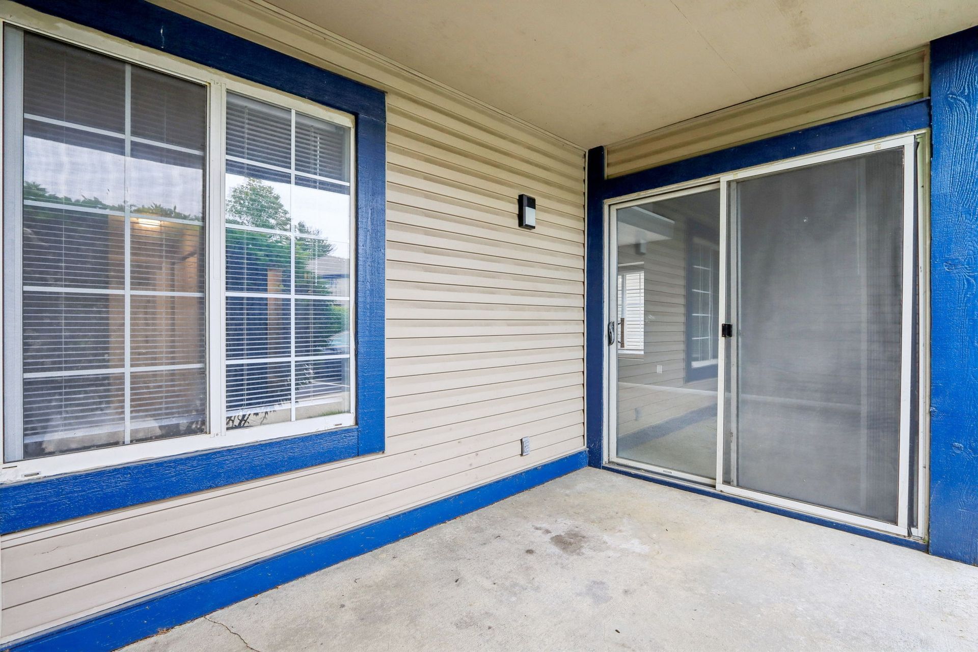 Covered patio with a sliding glass door and a window, blue trim, beige siding, and a concrete floor.