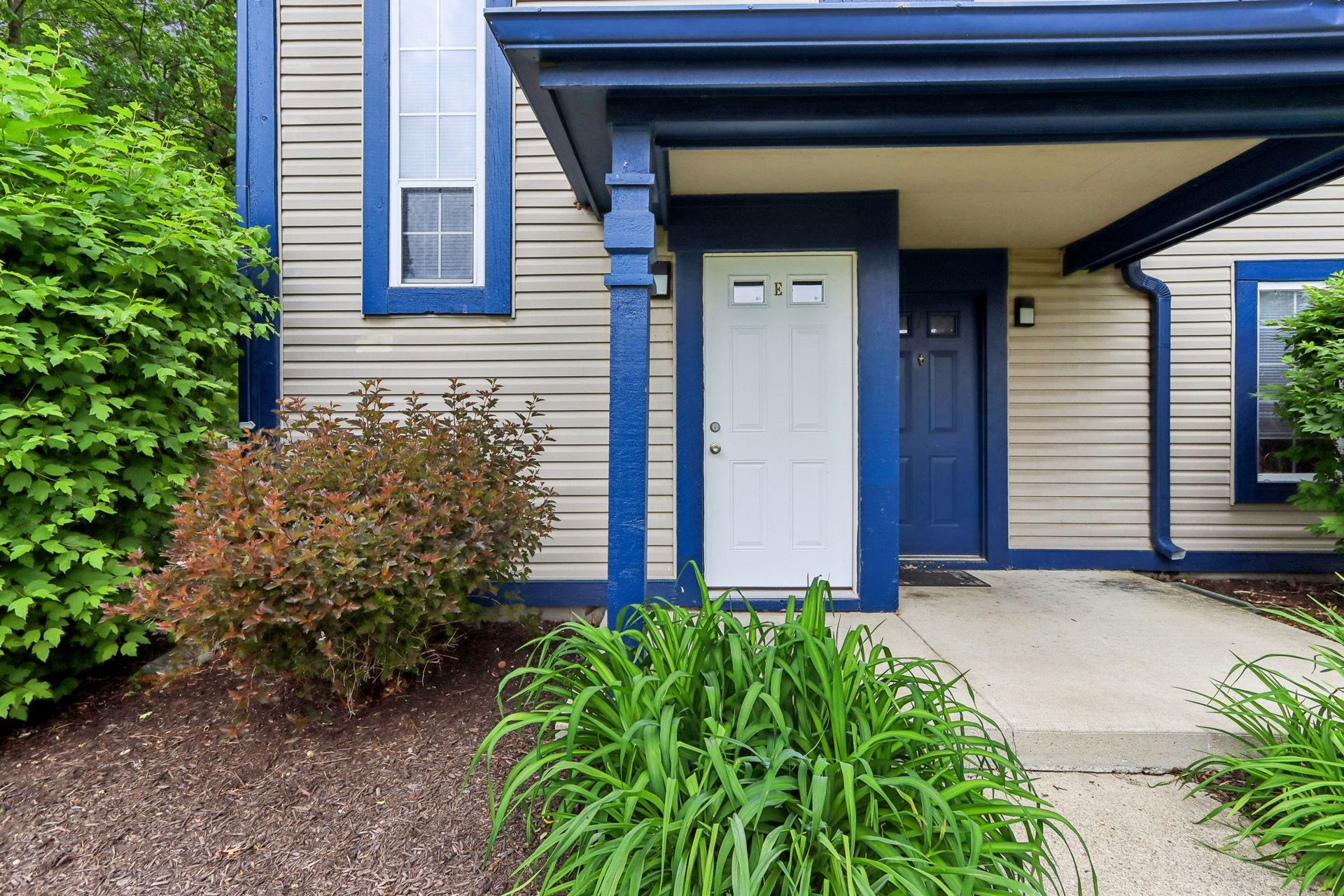 Exterior of a beige townhouse with blue trim and a white front door, surrounded by greenery.