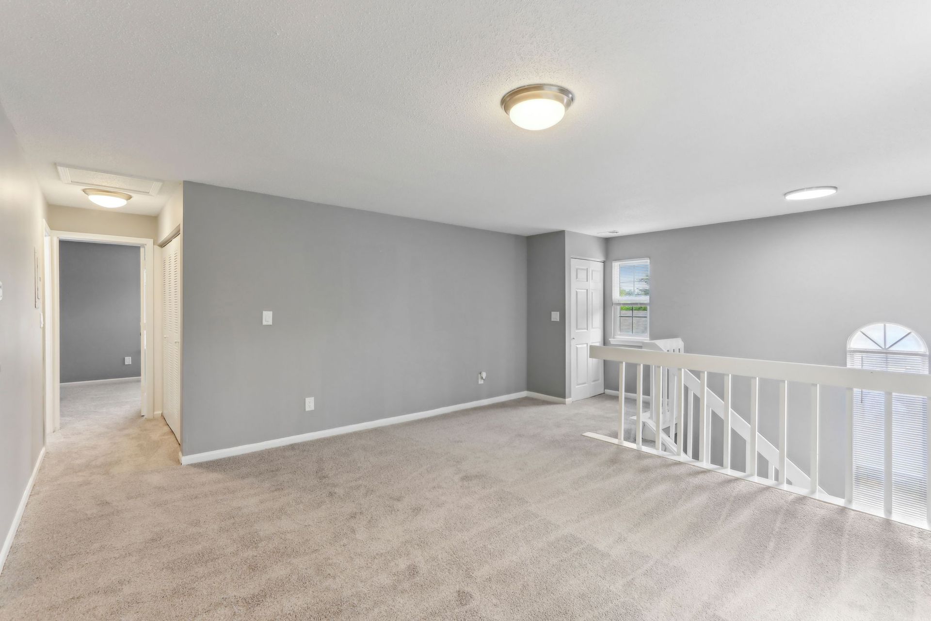 Empty gray-walled loft with carpet, white railing, and a hallway to the left.
