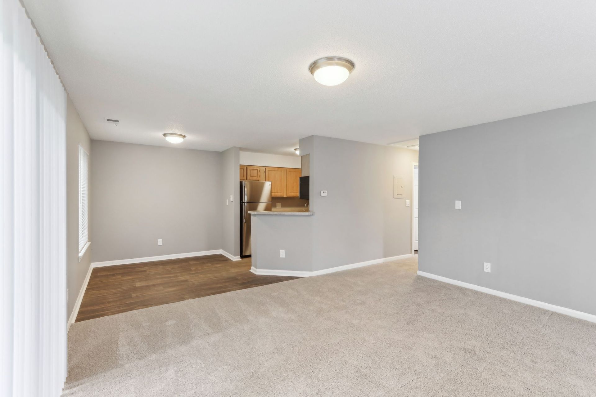 Empty living room with light gray walls, beige carpet, and kitchen in the background.