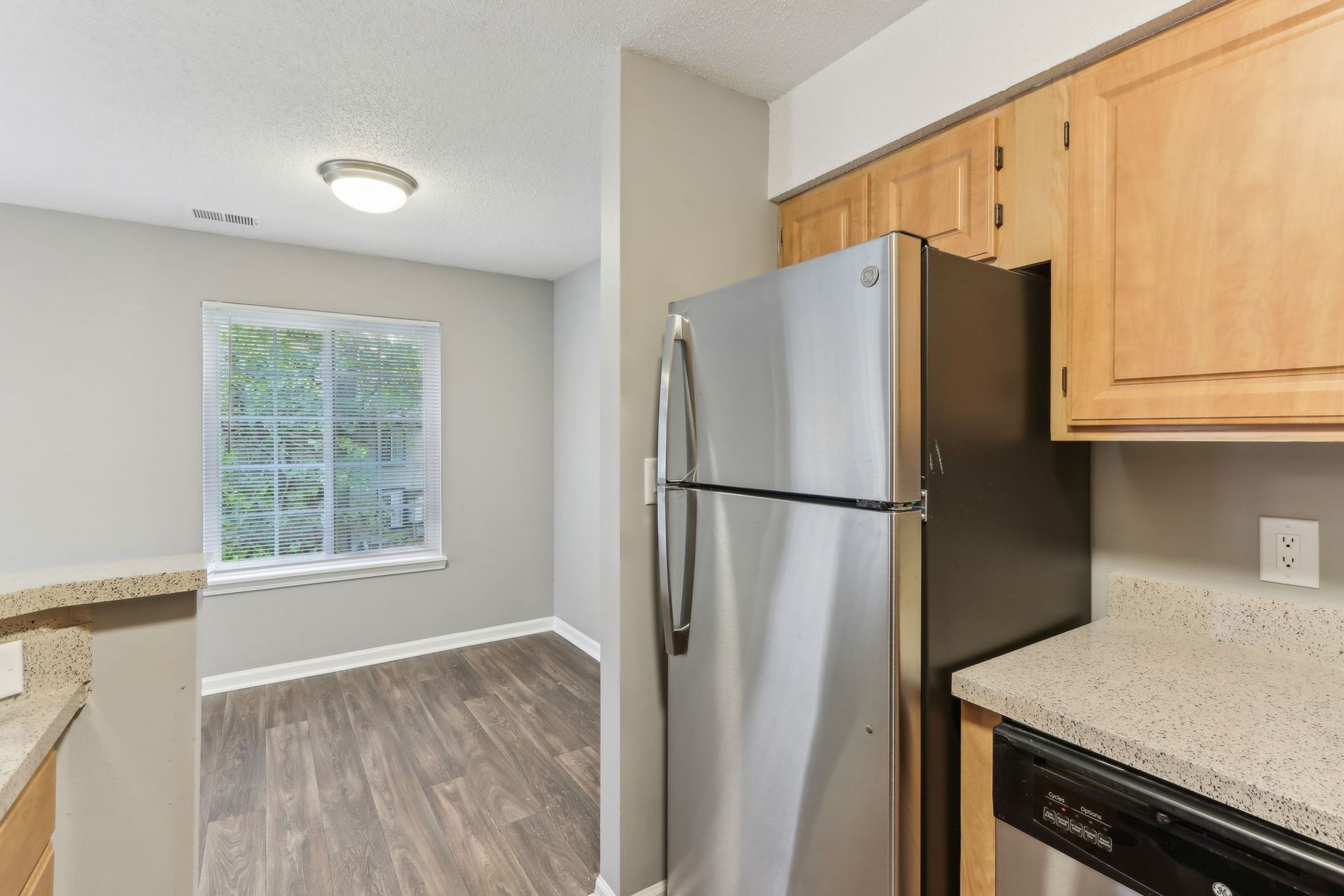 Kitchen with stainless steel refrigerator, wood cabinets, countertop, window, and wood flooring.