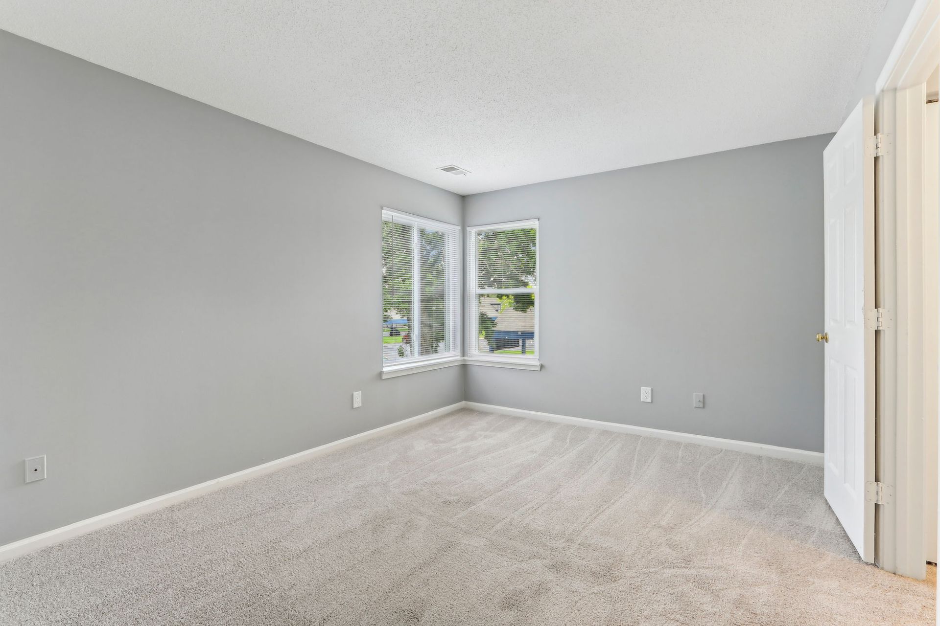 Empty bedroom with gray walls, carpet, and corner windows. A door is on the right side.