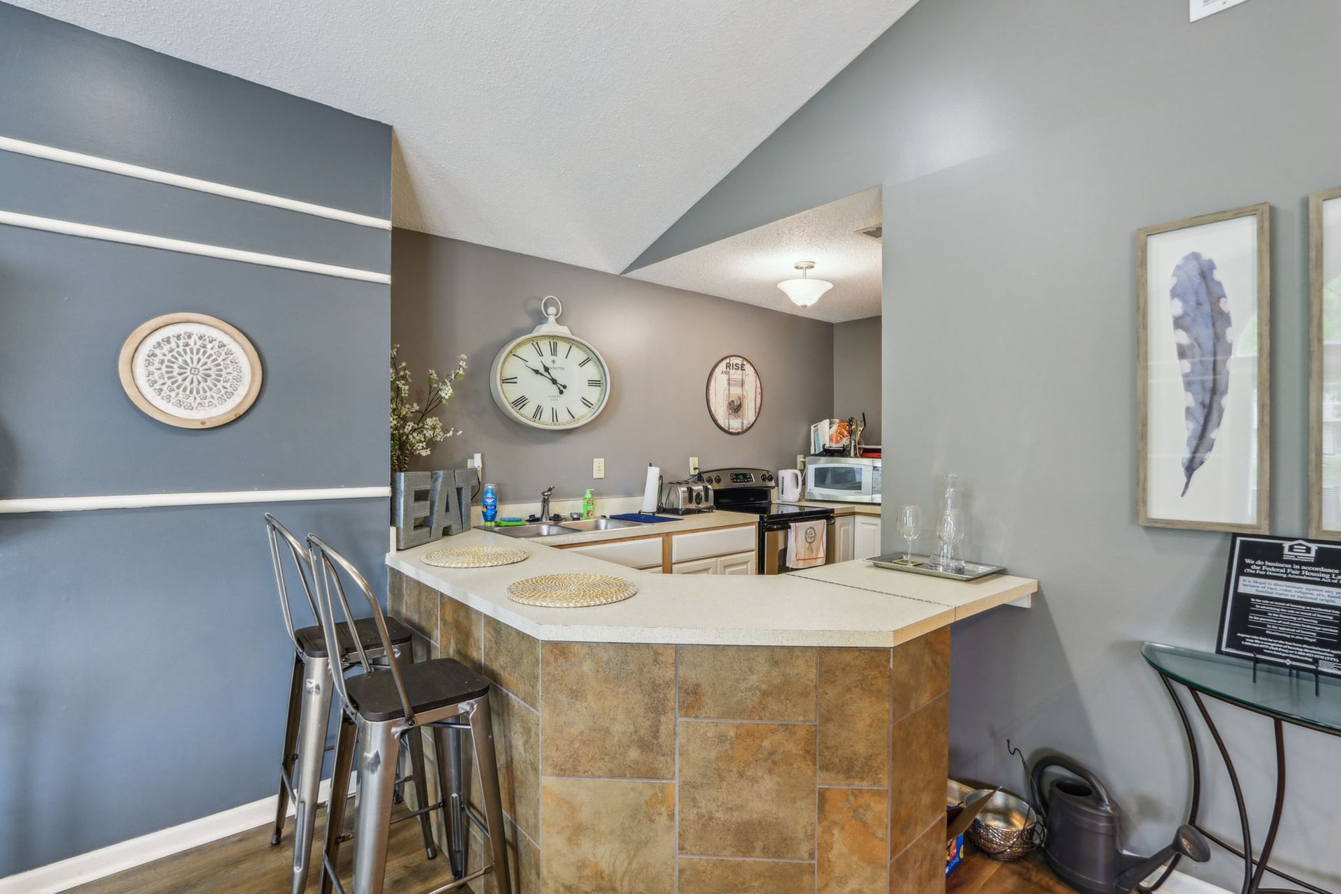 Kitchen with bar, stools, and decor. Gray walls, clock, sink visible in background.