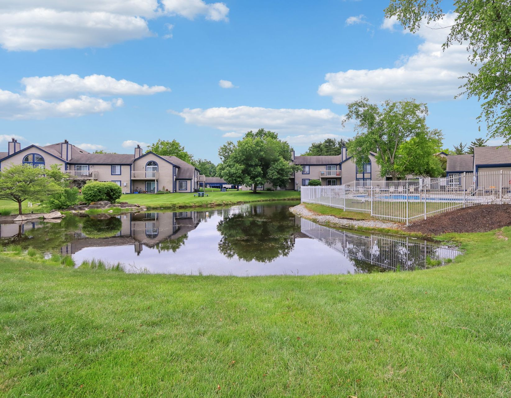Apartment complex with pond and pool on a sunny day, blue sky, green grass.