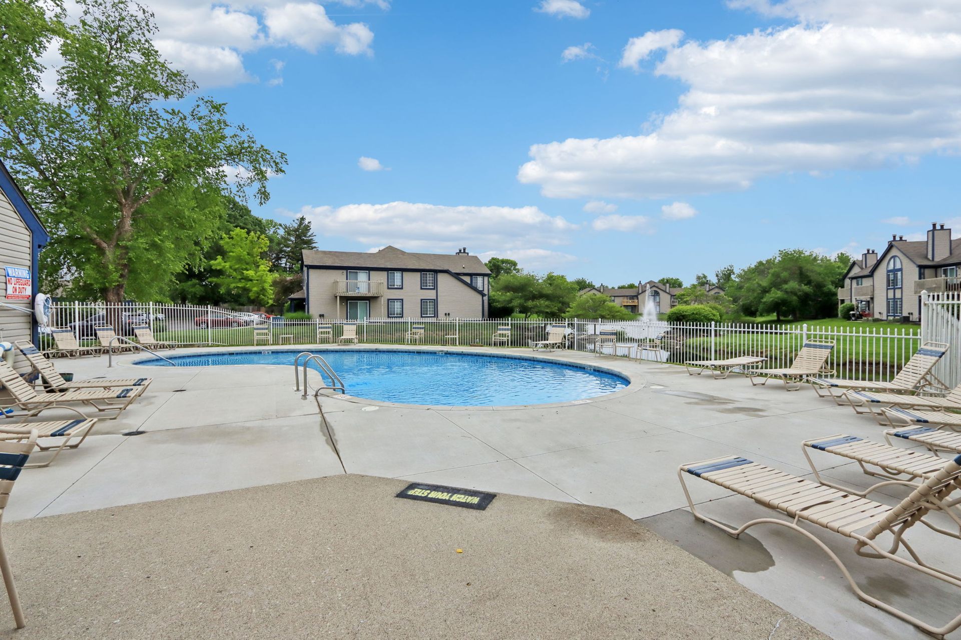 Pool area with a blue pool, lounge chairs, and apartment buildings under a cloudy sky.