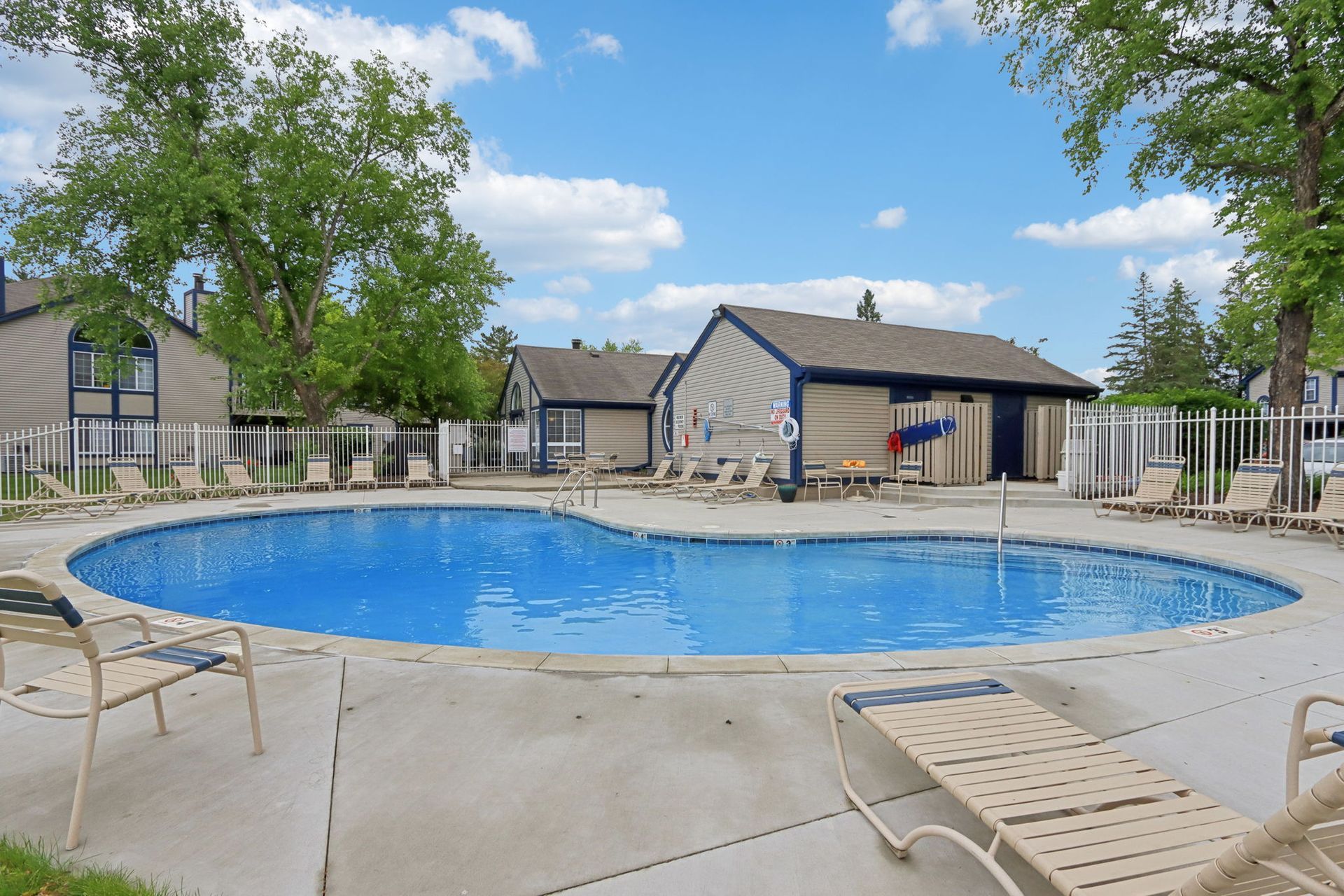 Swimming pool with lounge chairs, surrounded by beige buildings and a white fence, under a blue sky.