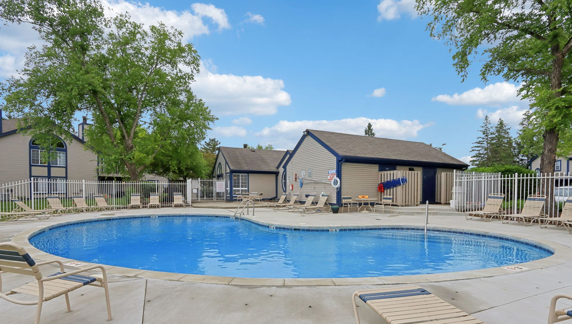 Swimming pool with lounge chairs, surrounded by buildings and trees under a blue sky.