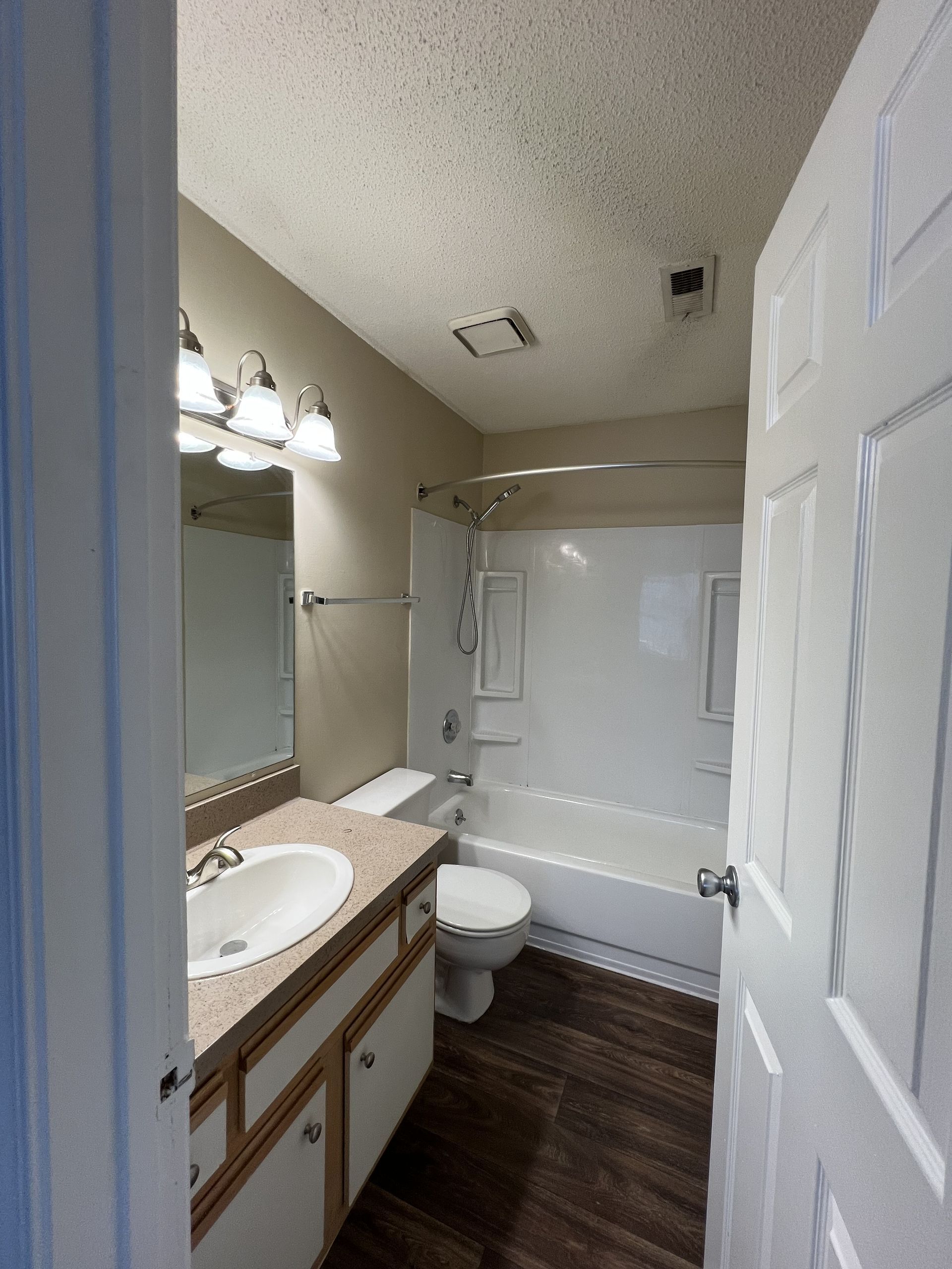 Bathroom with white tub, toilet, and vanity. Brown wood-look flooring and beige walls.