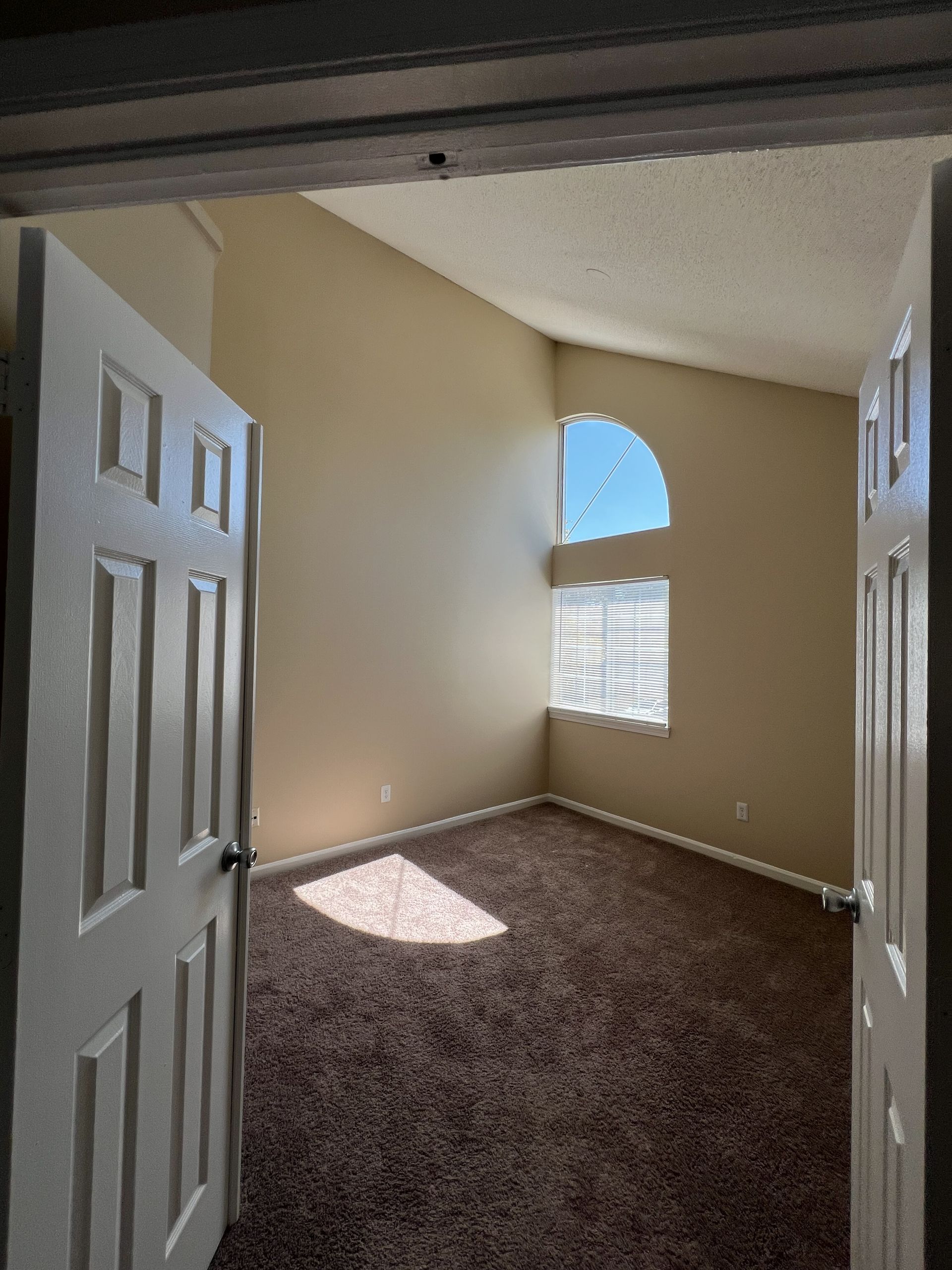 Empty room with brown carpet, tan walls, arched window, and open double doors.