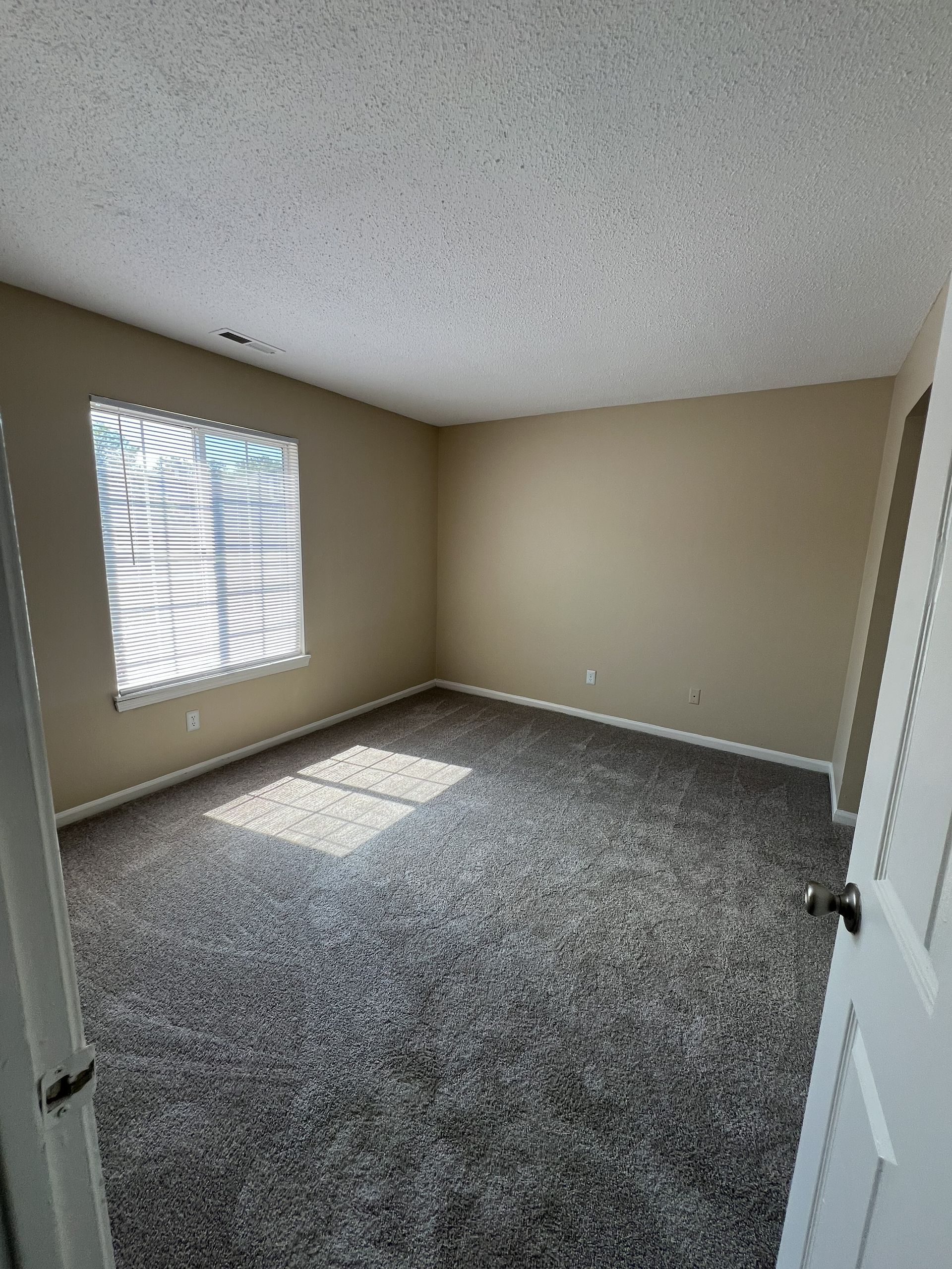 Empty bedroom with neutral walls, gray carpet, and a window letting in sunlight.