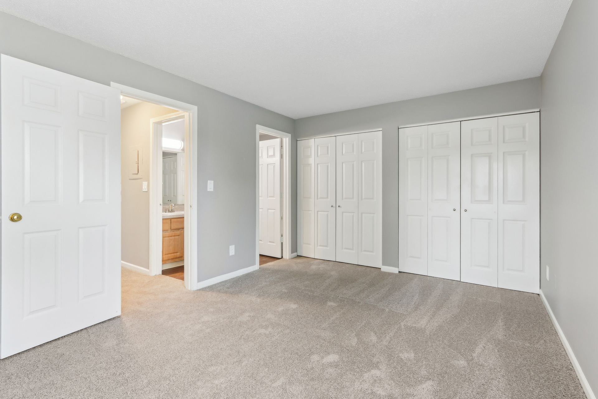 Bedroom with gray walls, white doors, and beige carpet. Two closet doors on the right, open bathroom door to the left.