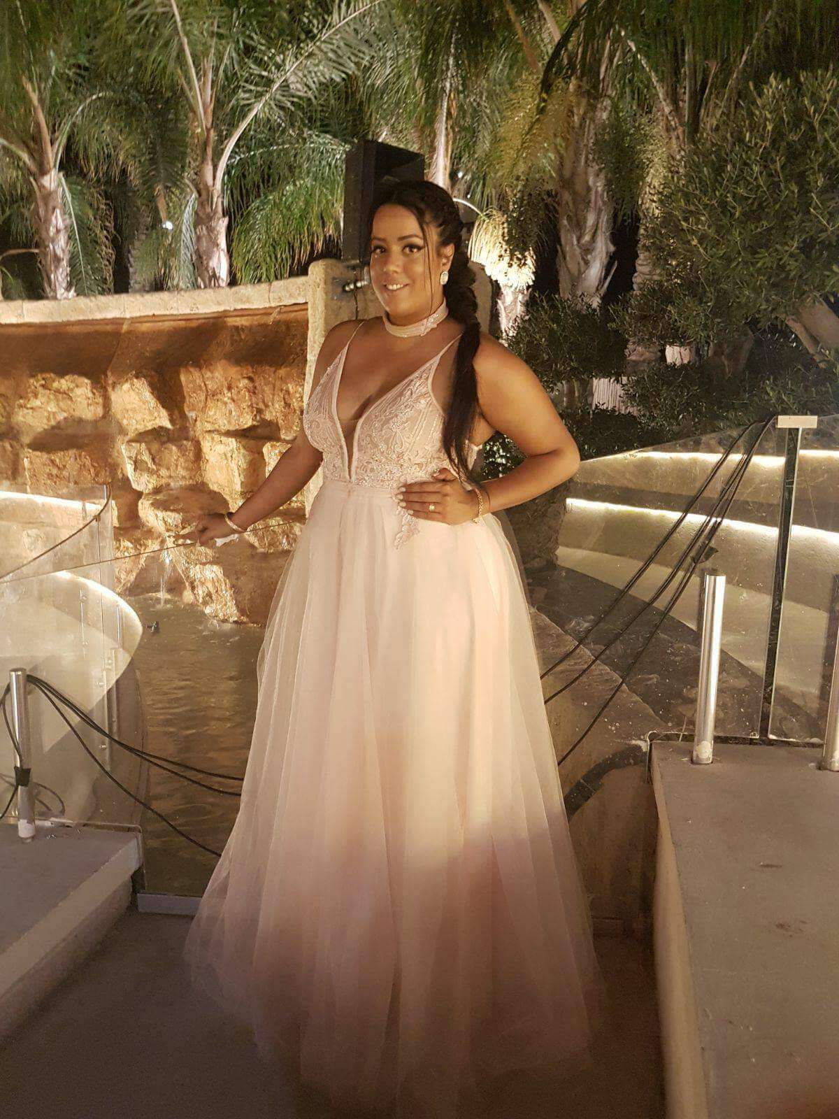 אישה בשמלה רשמית ורודה בהיר עומדת בחוץ, מחייכת, ליד מתקן מים וירק.