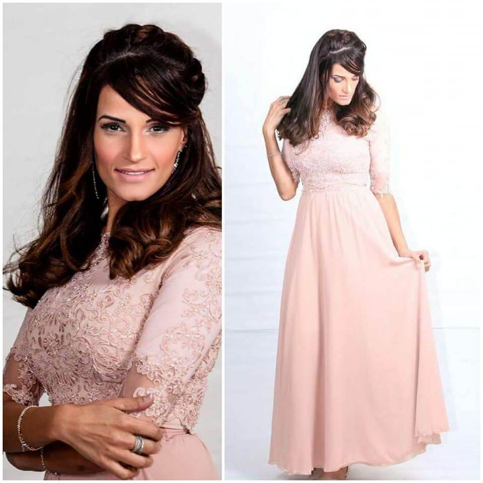 אישה בשמלת תחרה ורודה מצטלמת, מביטה למטה, עם שיער חום ארוך אסוף.