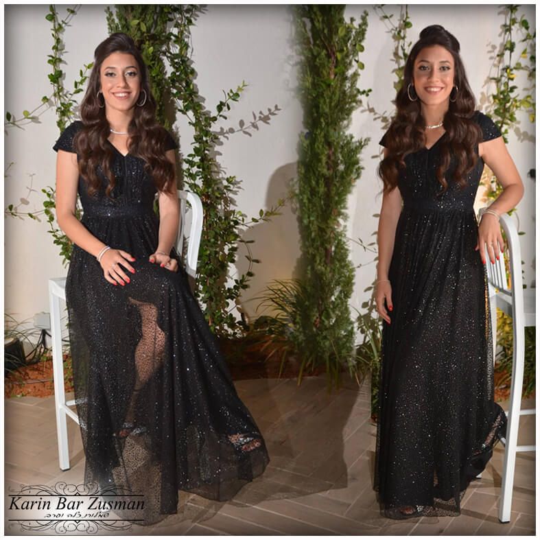 אישה בשמלה שחורה, מחייכת, מצטלמת על כיסא לבן. עלים ירוקים עבותים ברקע.